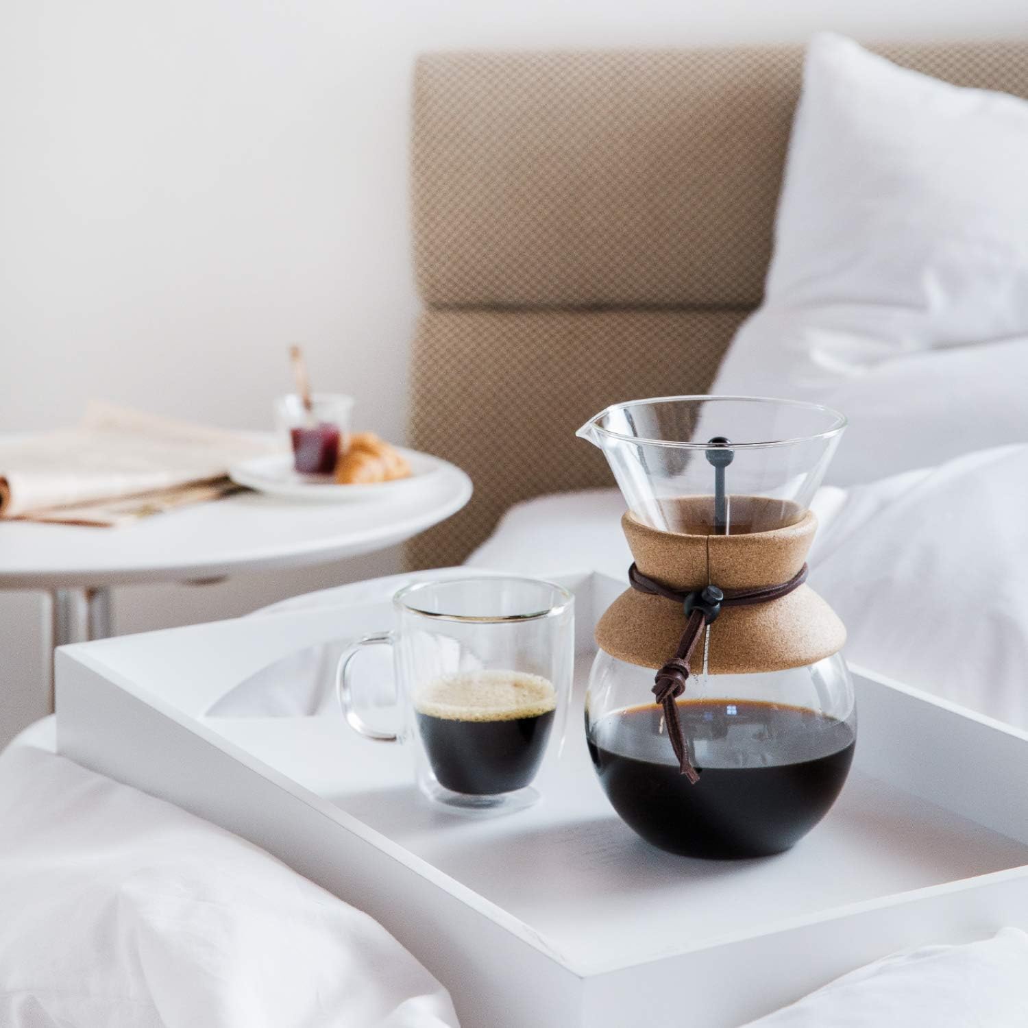 Glass pour-over coffee maker with a cork grip and leather tie on a white tray beside a cup of coffee